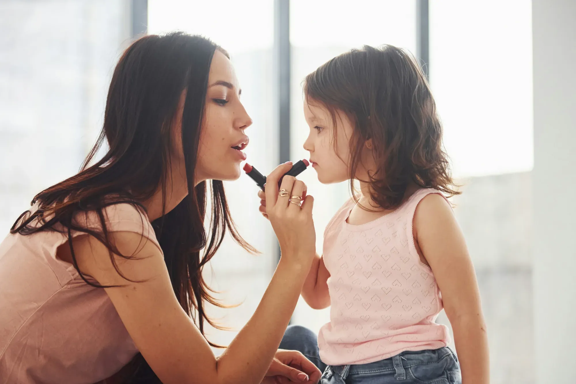 A mother putting on lipstick beside her young daughter in a bright window-lit room as the girl watches closely