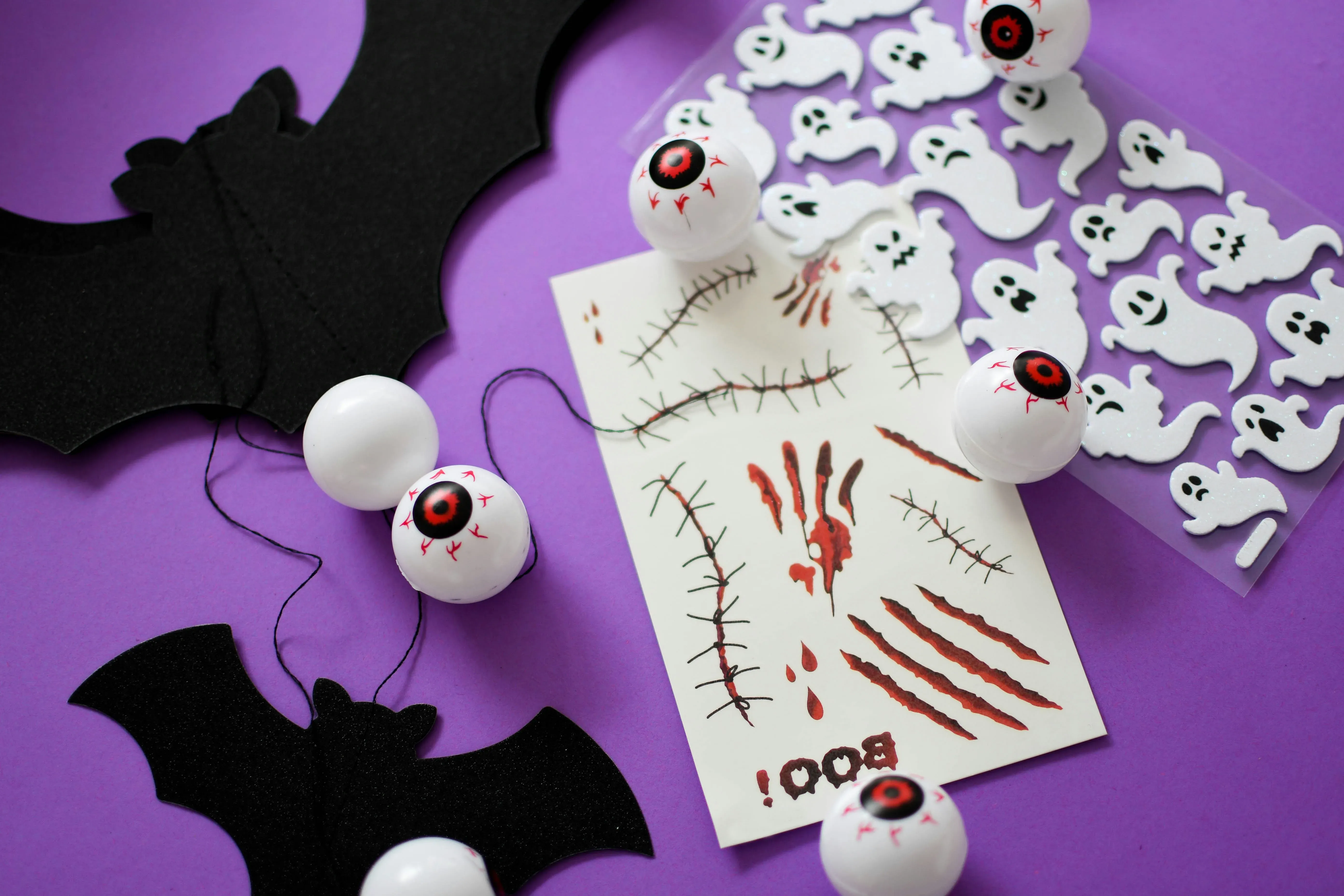 Halloween party favors on a purple background: black paper bats, plastic eyeballs, a sheet of glitter ghost stickers and a sheet of spooky temporary tattoos