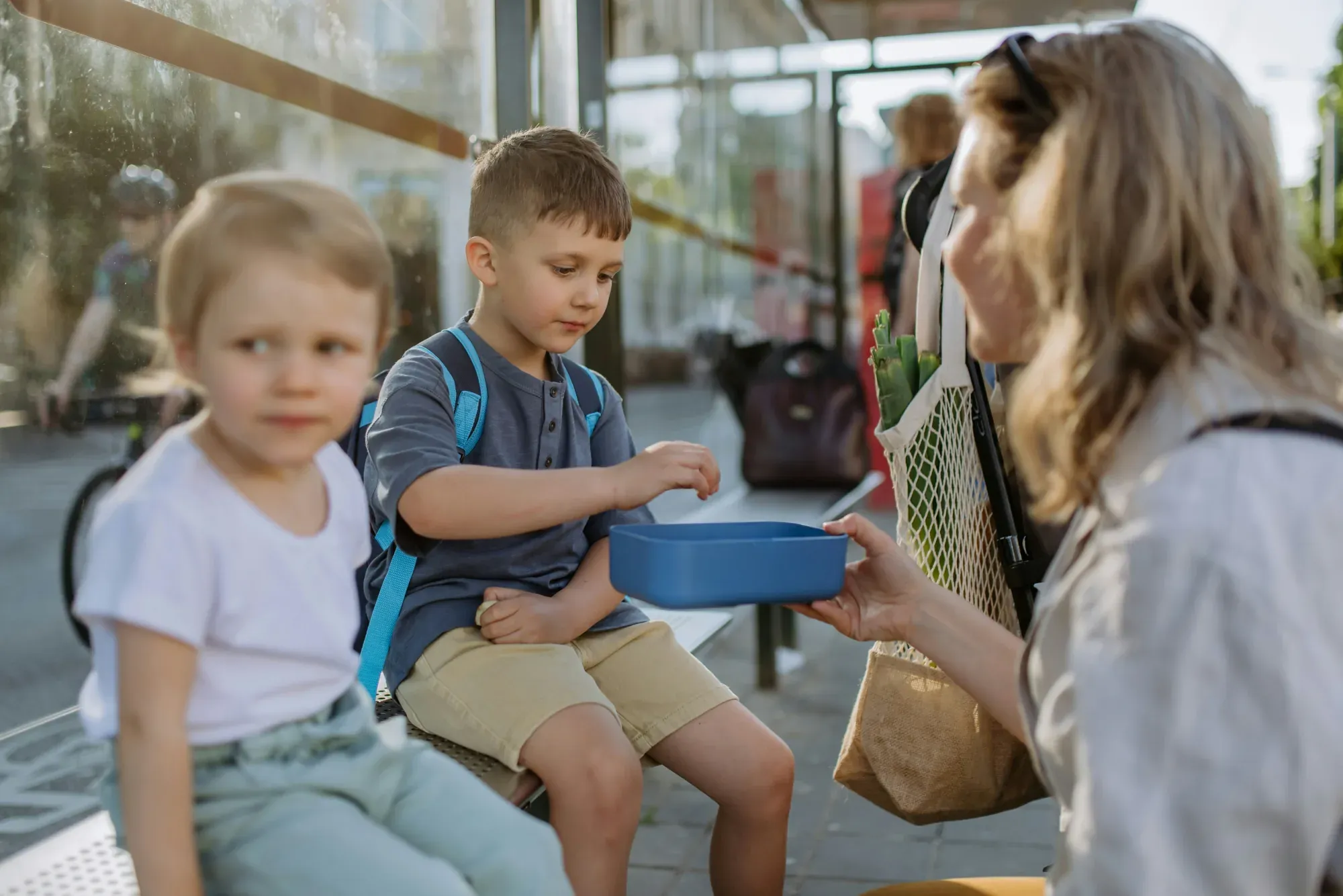 A mother handing a blue lunchbox to her young son on a bus stop bench while his little sister waits beside him