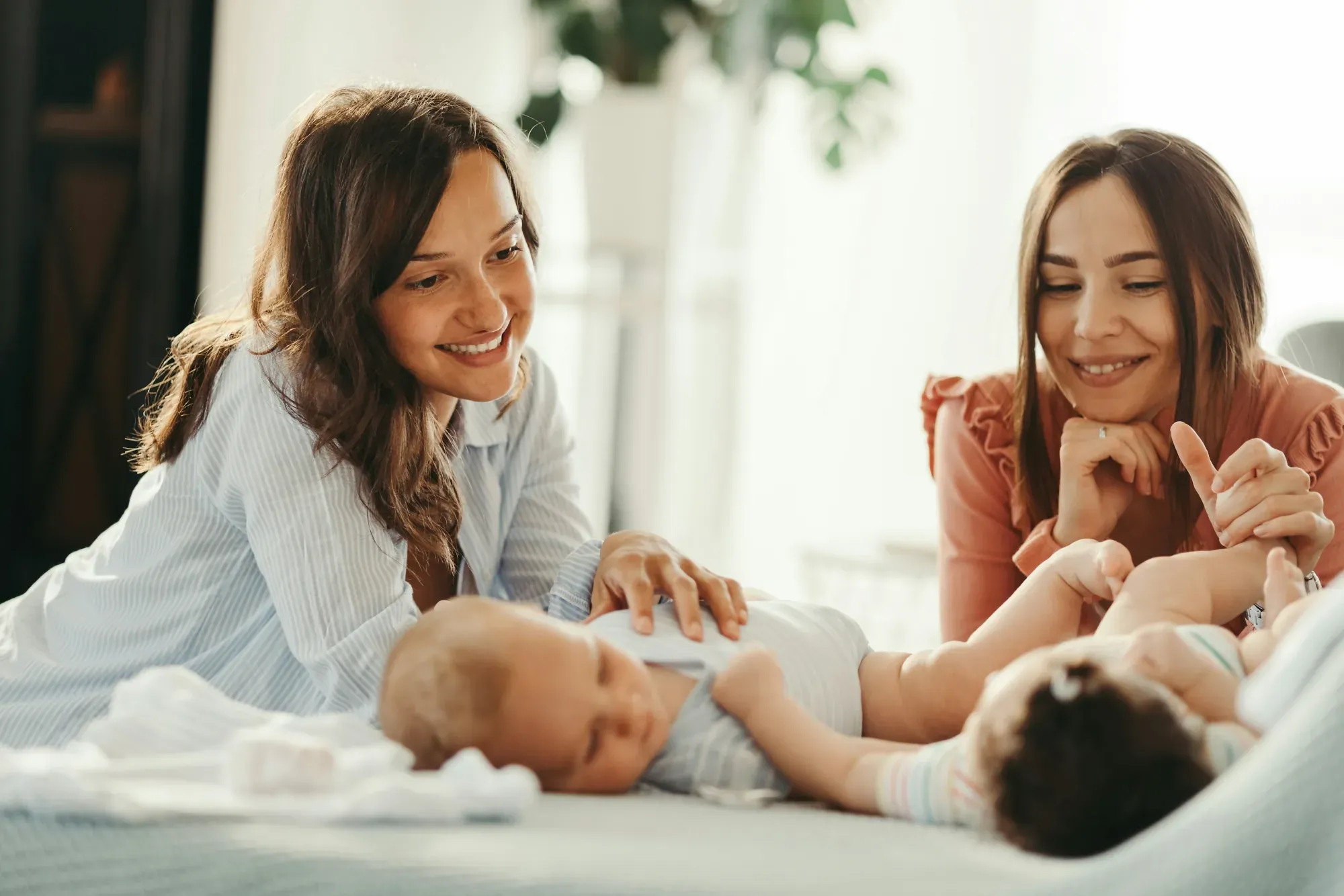 Two mothers leaning over their babies on a bed in a sunlit room, smiling as they play with them