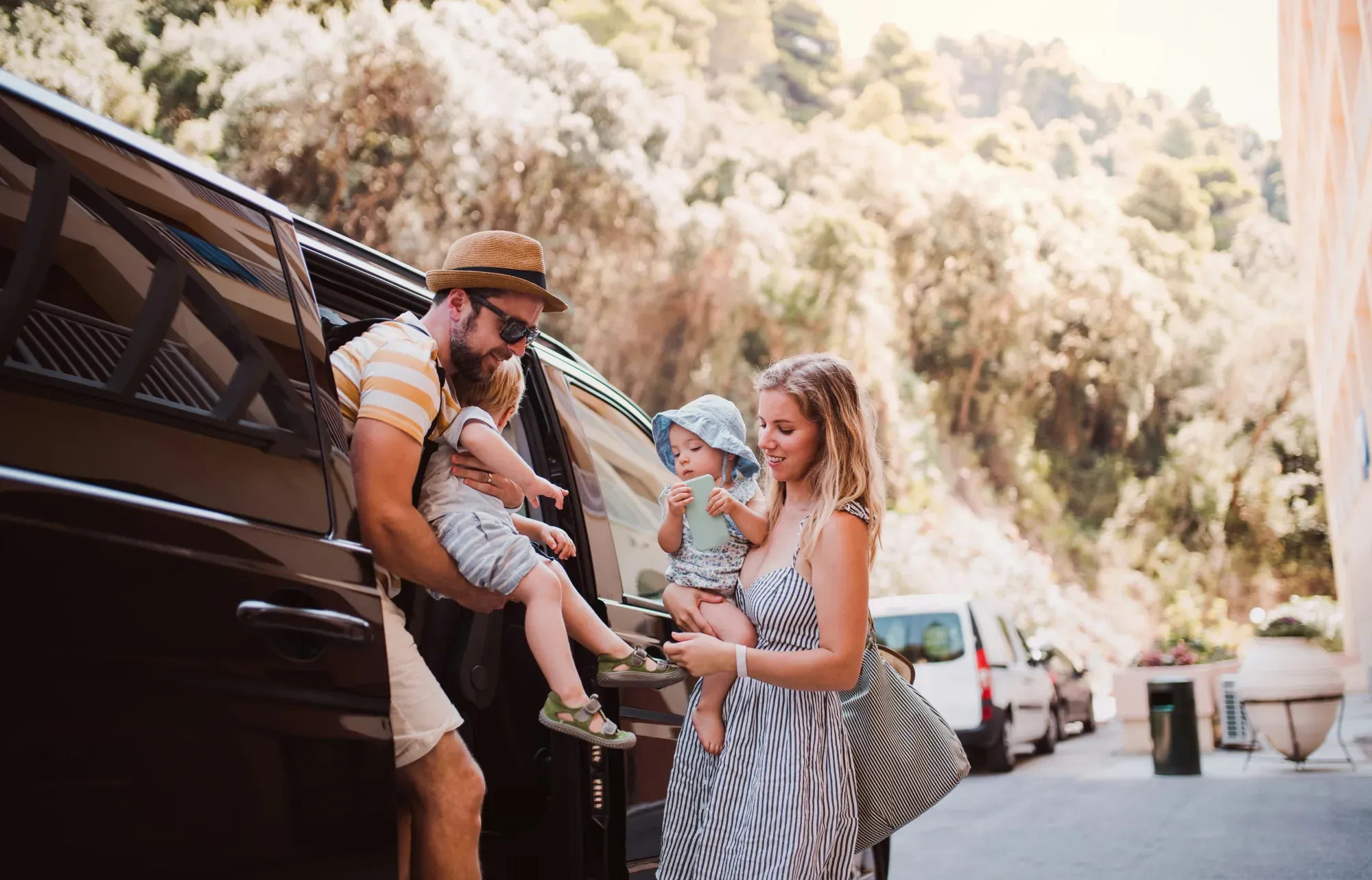 Parents lifting their two toddlers out of a van on a sunny tree-lined street at the start of a weekend trip