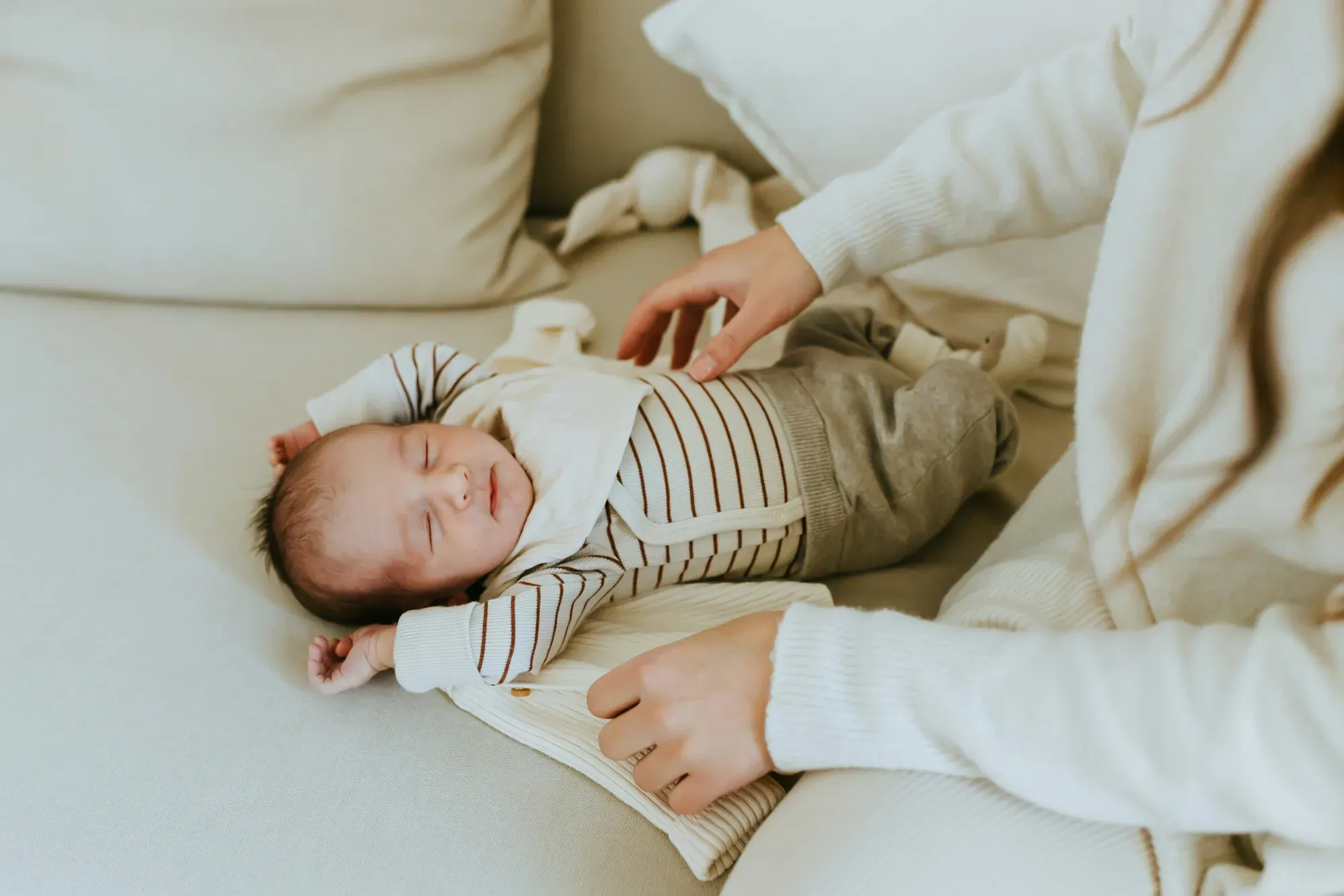 A mother's hands resting gently on her sleeping newborn, who is dressed in striped cream clothes on a soft neutral couch