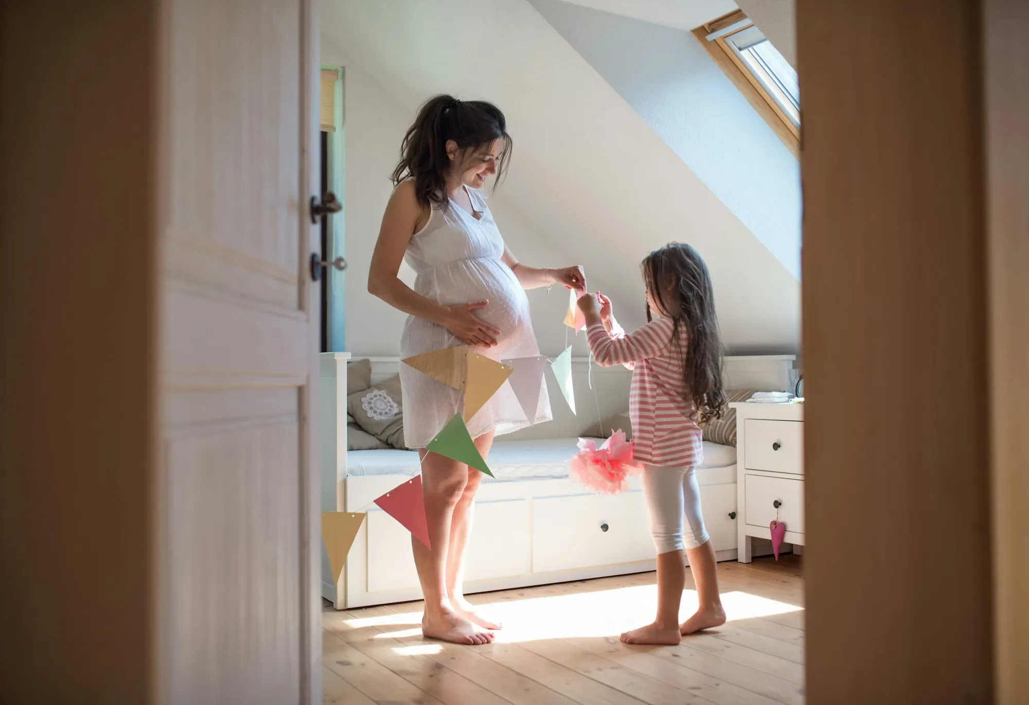 A pregnant woman and her young daughter hanging a paper bunting garland together in a sunlit attic bedroom