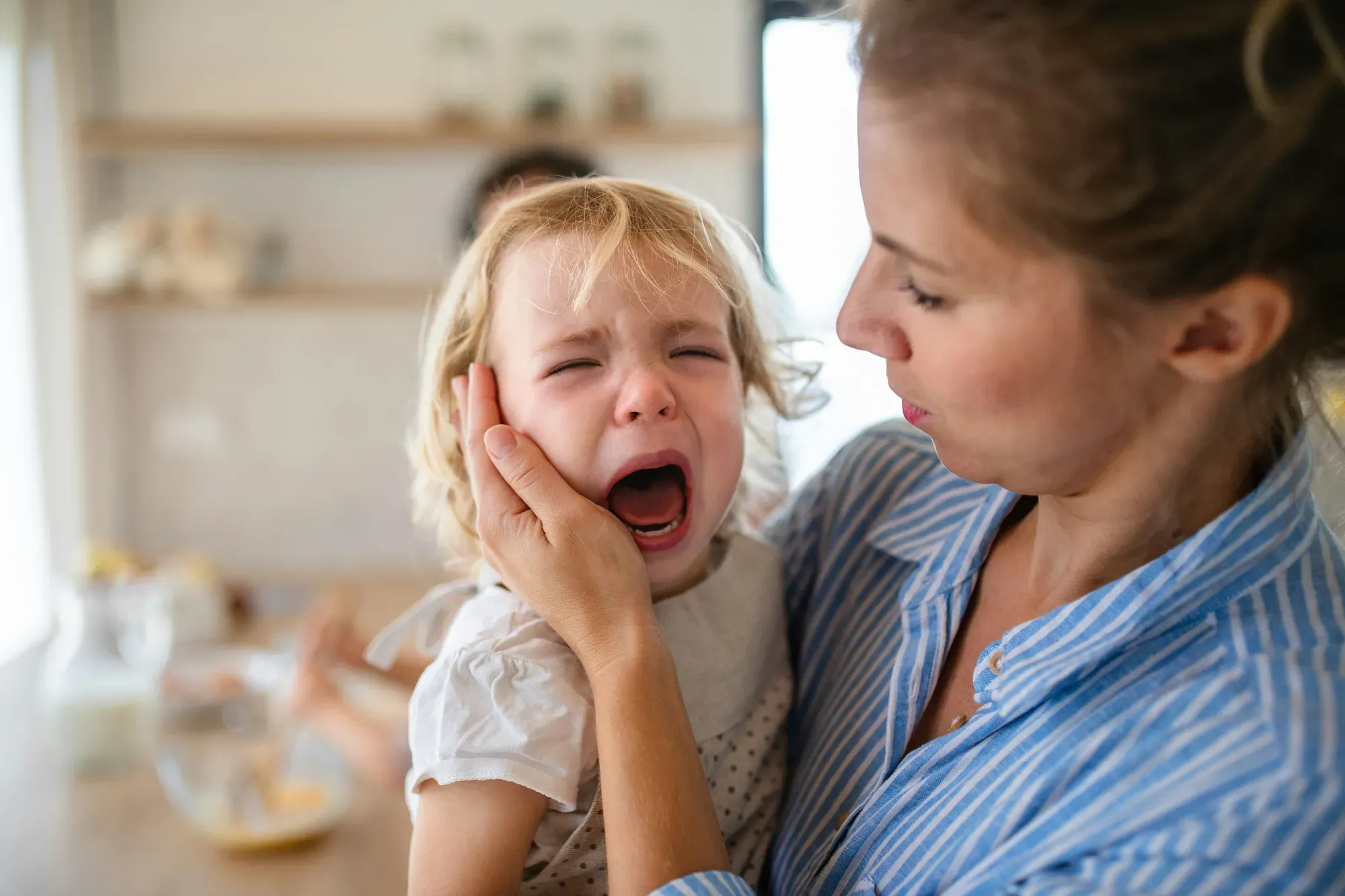 A mother in a blue striped shirt holds her crying toddler and cradles the little girl’s cheek in a sunny kitchen